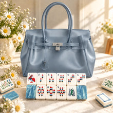 Gray-blue structured handbag displayed behind red-and-blue Mahjong bamboo tiles with blue marbled backs and white daisies.