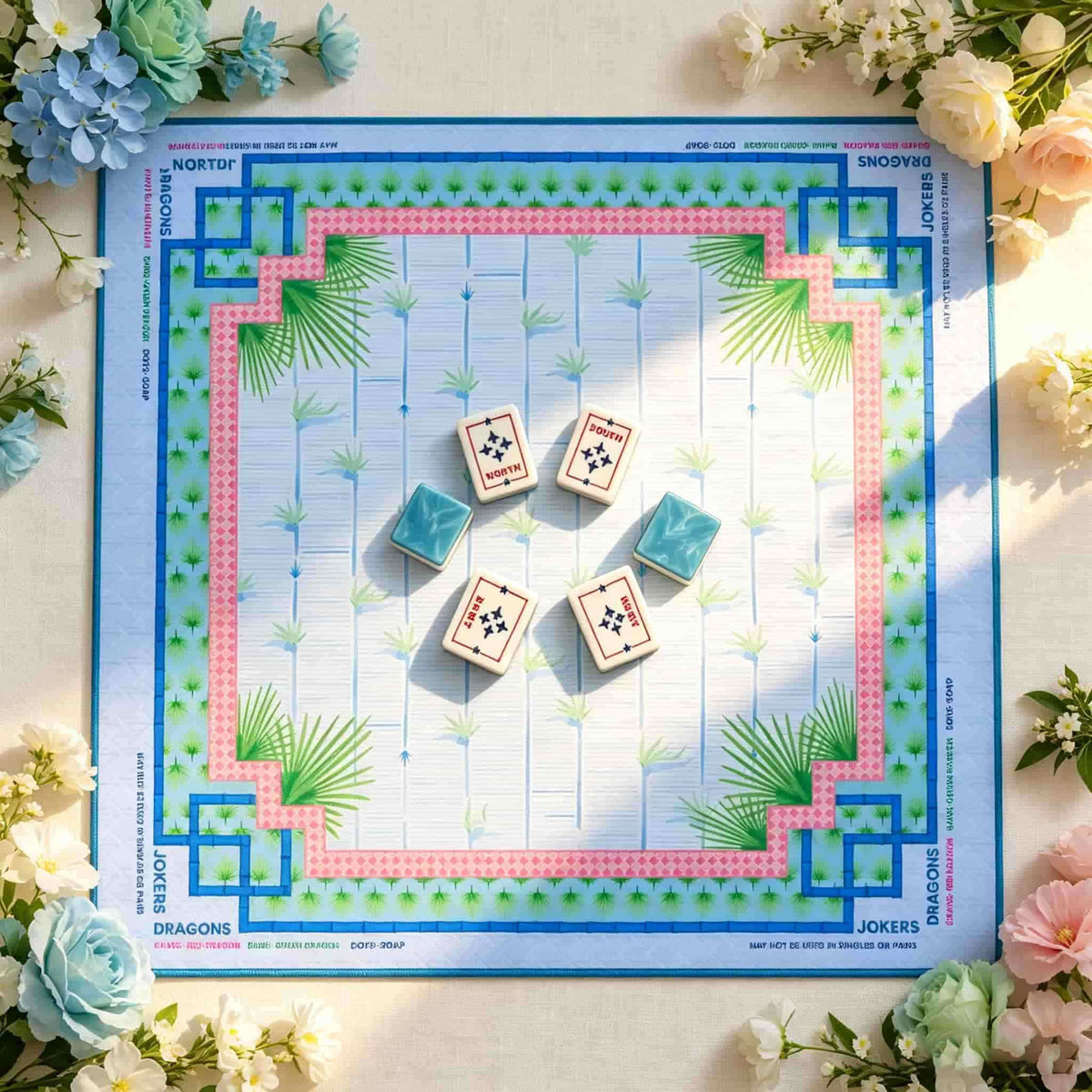 Square blue Mahjong mat with a pink border, palm fronds and geometric corners, shown with wind tiles and blue-backed tiles.